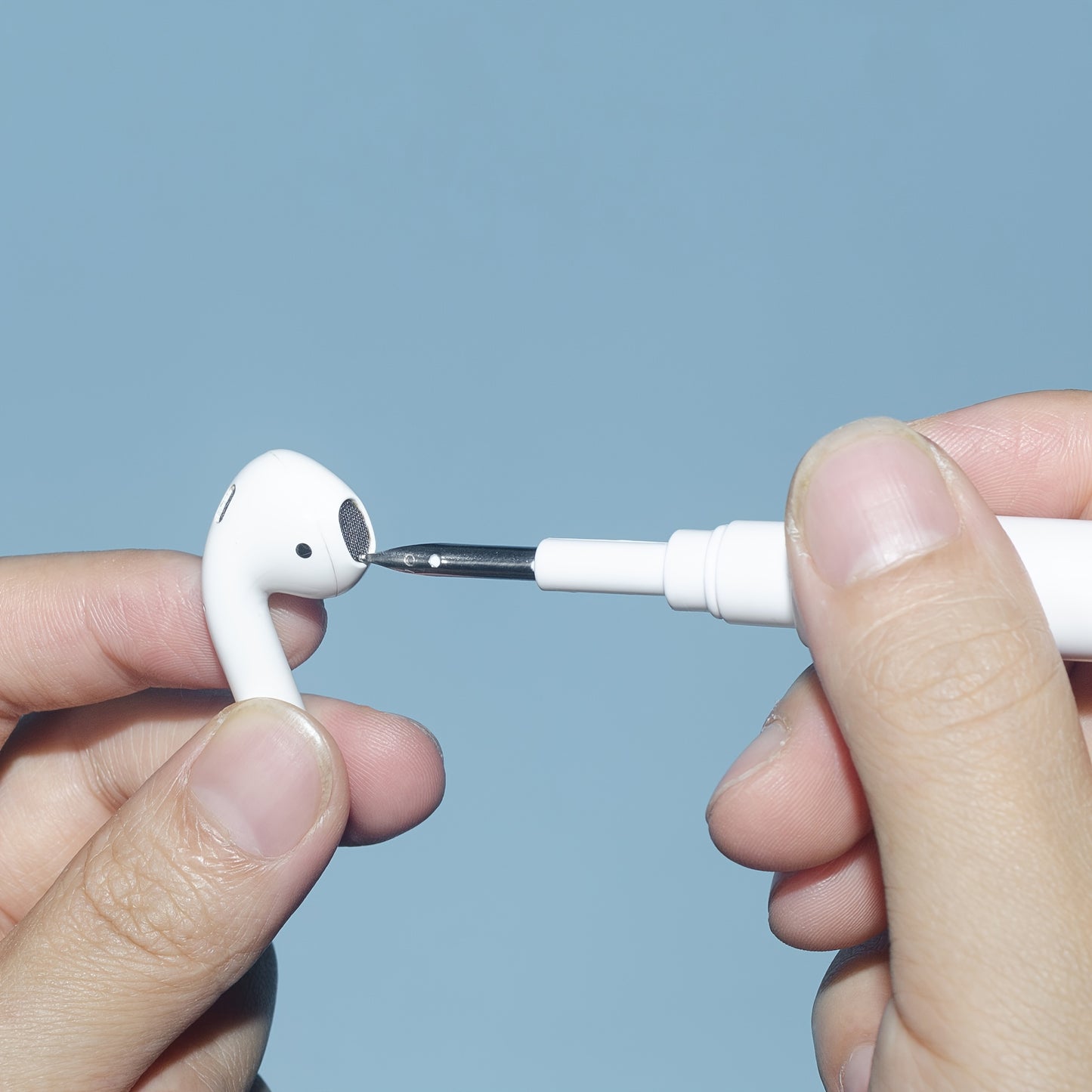Person cleaning a white wireless earbud with a small brush against a light blue background