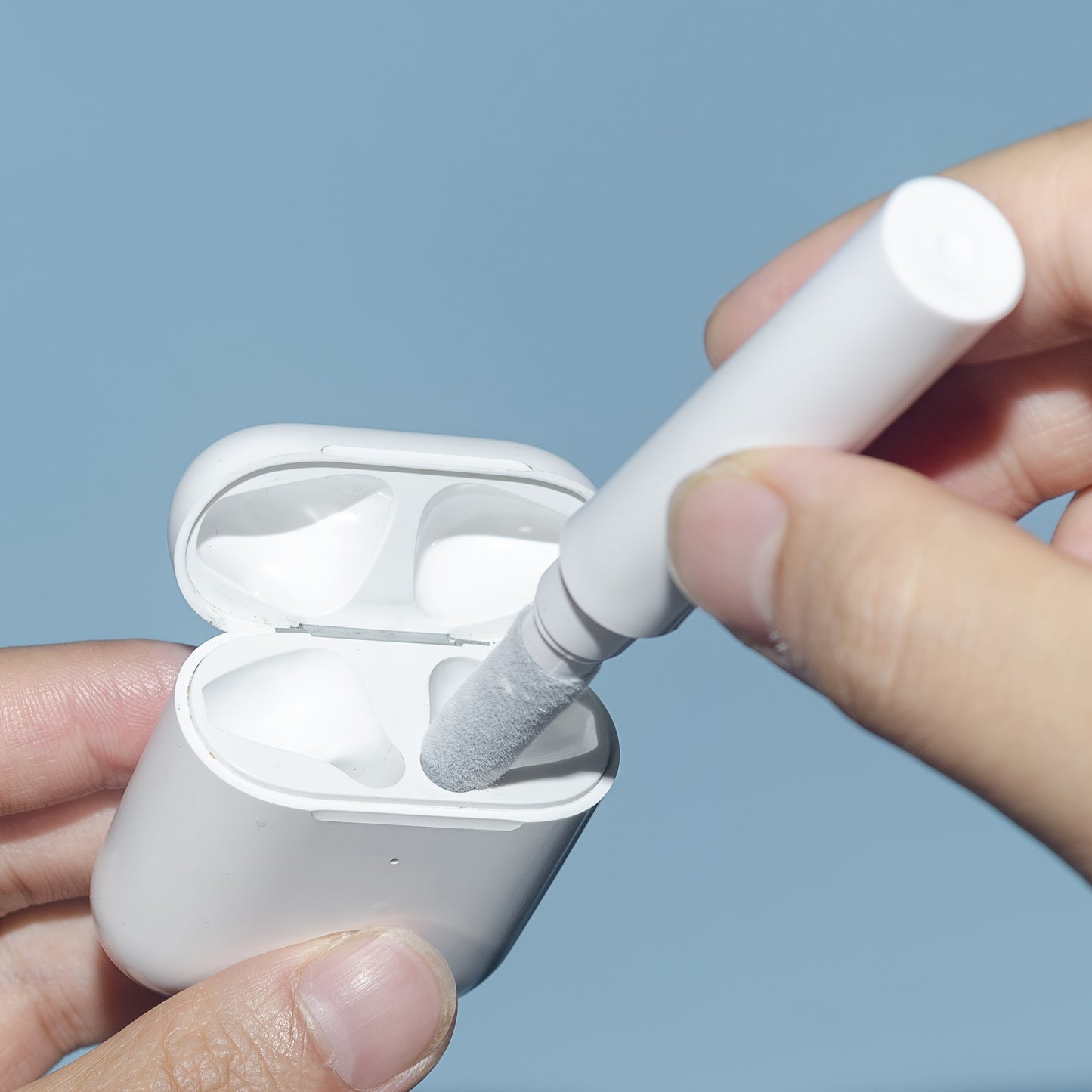 Person cleaning white earbuds with a small brush against a light blue background