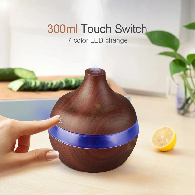 Wooden diffuser with blue LED light on a table with a hand touching the switch, surrounded by cucumbers and a lemon.