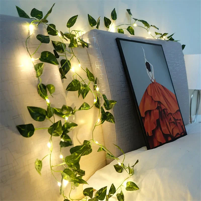 Decorative string lights with green leaves draped over a bookshelf with a painting on it.