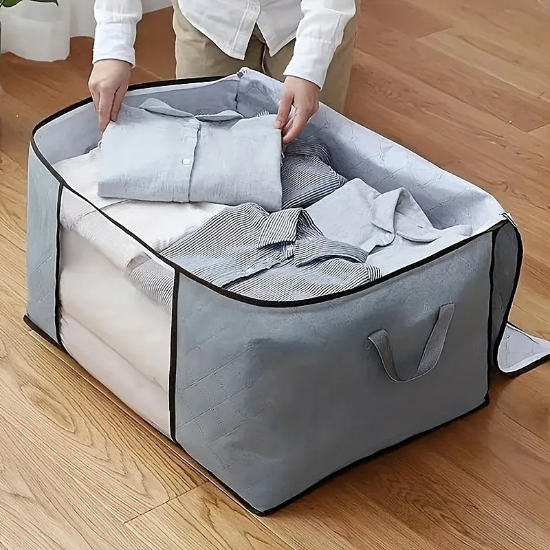 Person organizing folded clothes into a large gray storage bag on a wooden floor.