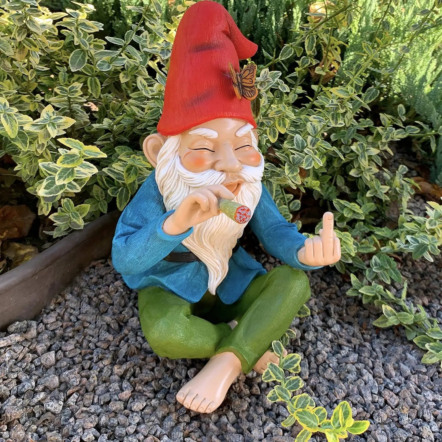 Gnome figurine with red hat and blue coat sitting on gravel with green plants in the background