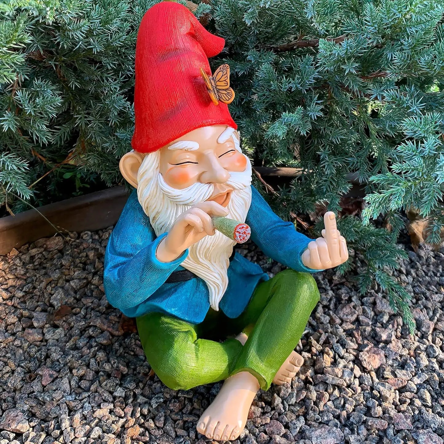 Gnome figurine with a red hat and green pants sitting on gravel with plants in the background