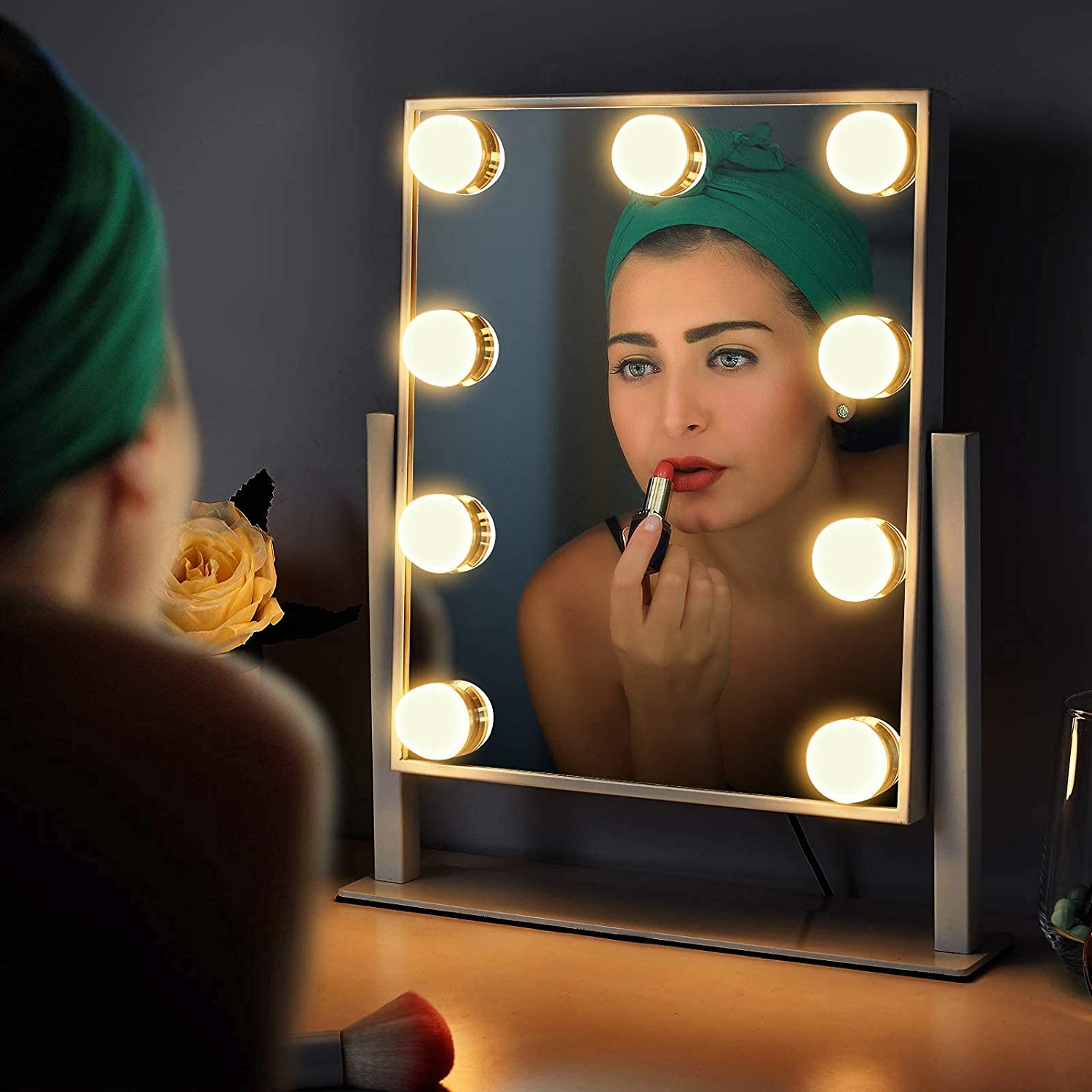 Woman applying makeup in front of a mirror with lights on a dark background
