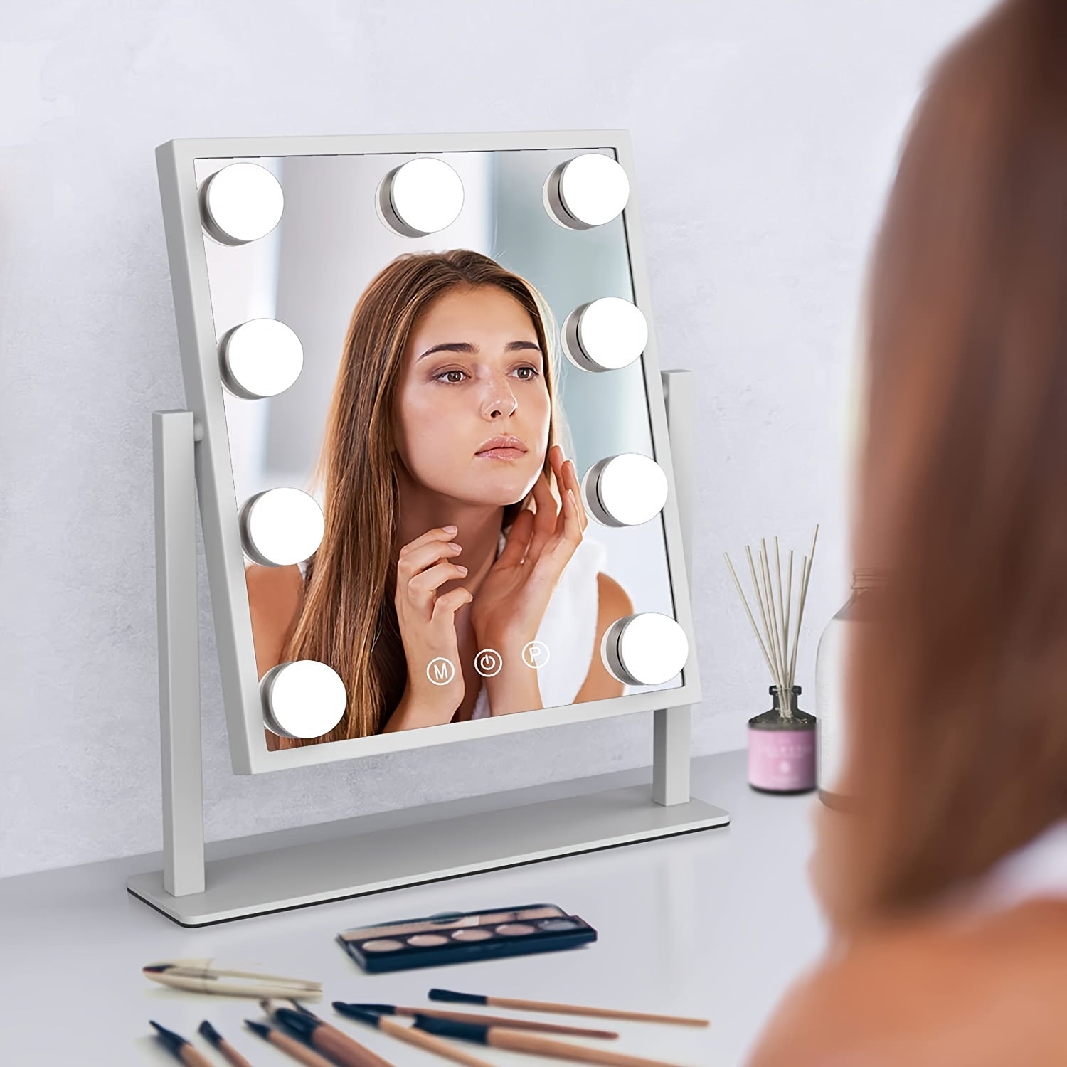 Woman using a vanity mirror with lights on a table with makeup items.