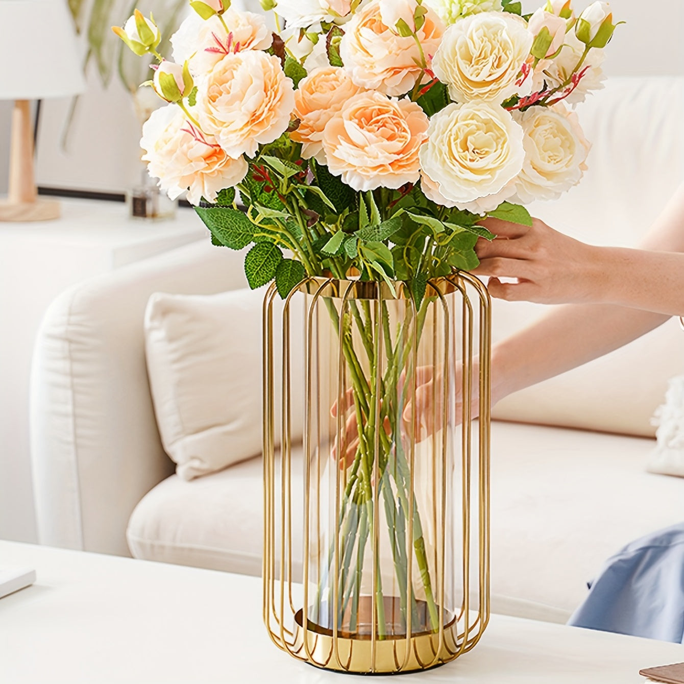 Person holding a bouquet of flowers in a gold vase on a table with a white couch in the background