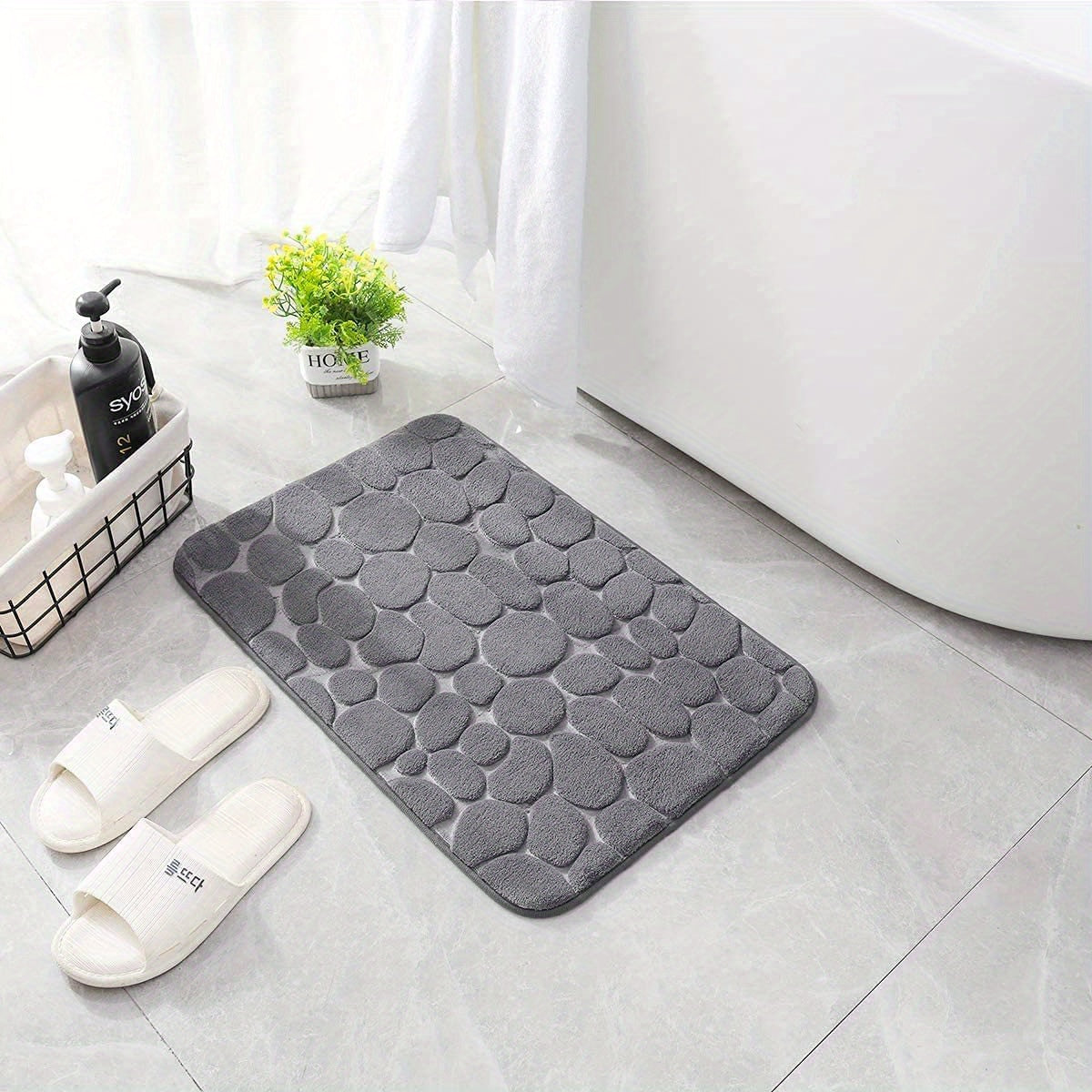 Gray textured bath mat on a bathroom floor with white slippers and a plant.