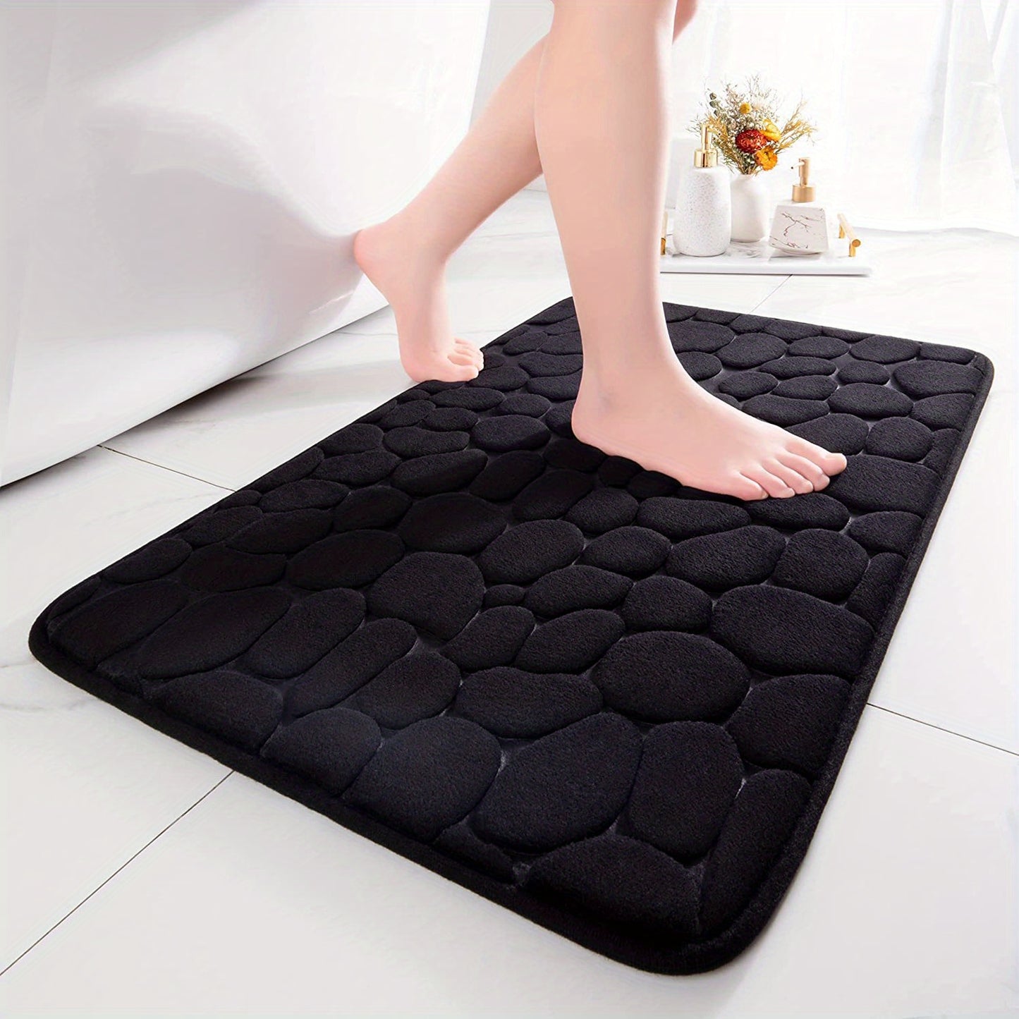 Black textured bath mat with feet stepping onto it on a white tiled floor.
