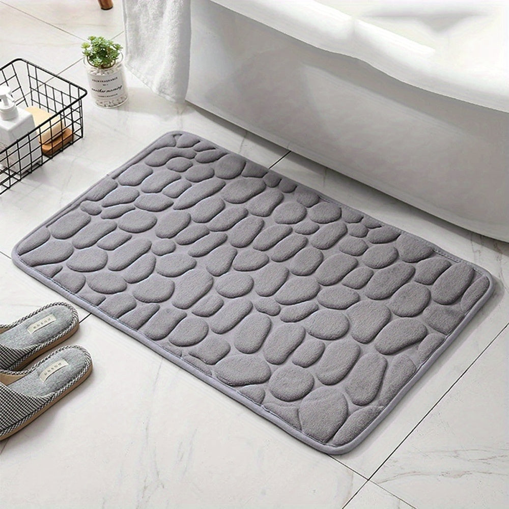 Gray textured bath mat on a bathroom floor with slippers and a plant in the background.