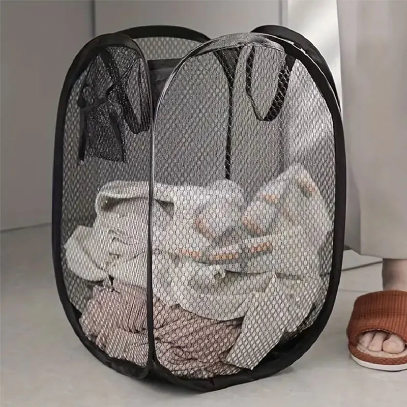 Black mesh laundry basket with clothes inside on a light-colored floor.