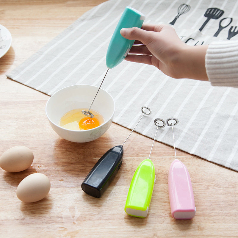 Hand using a turquoise handheld mixer to whisk eggs in a bowl on a wooden surface.