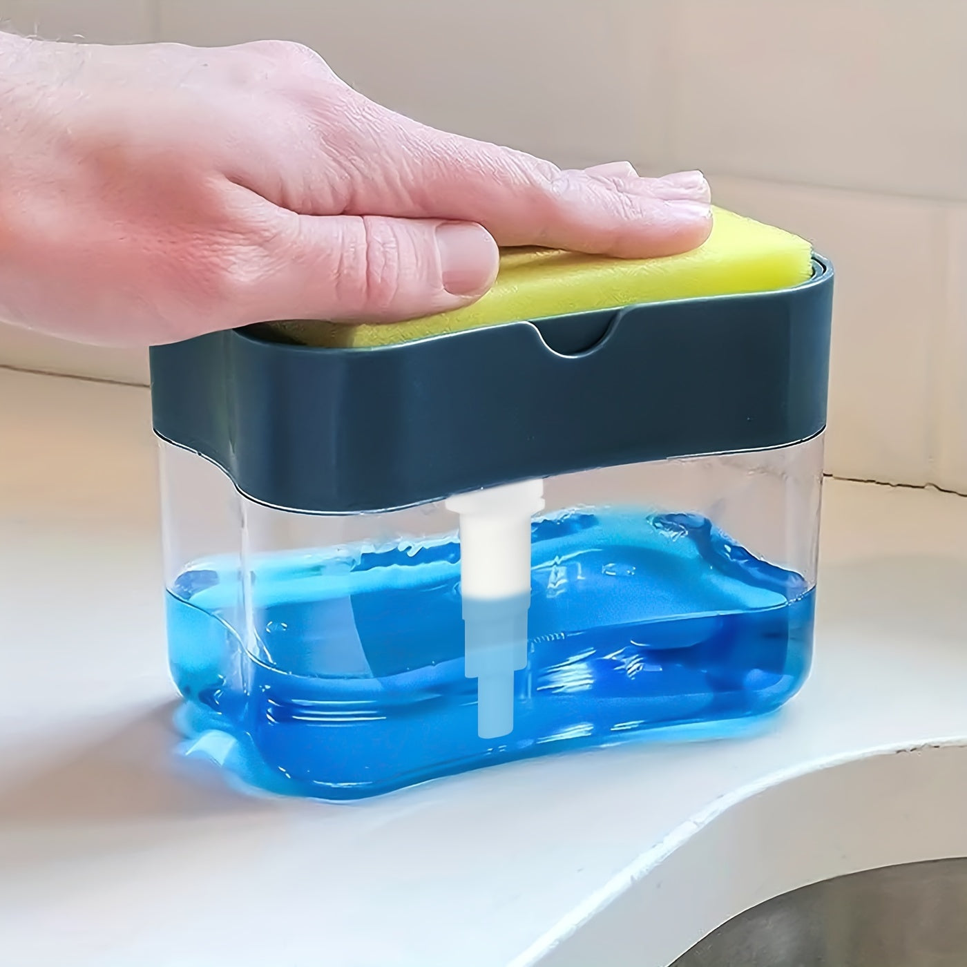 Hand holding a yellow sponge over a blue soap dispenser on a white surface.