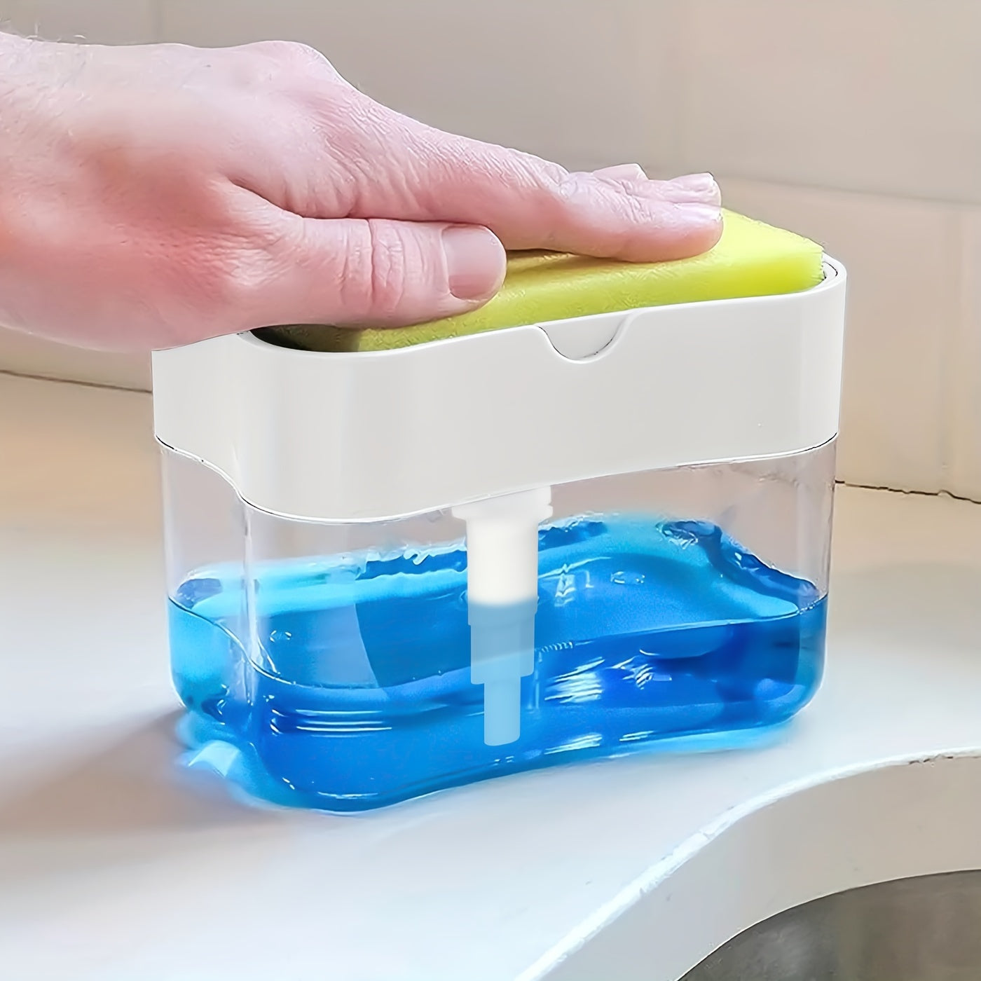Hand holding a green sponge over a white and clear container with blue liquid on a light surface.