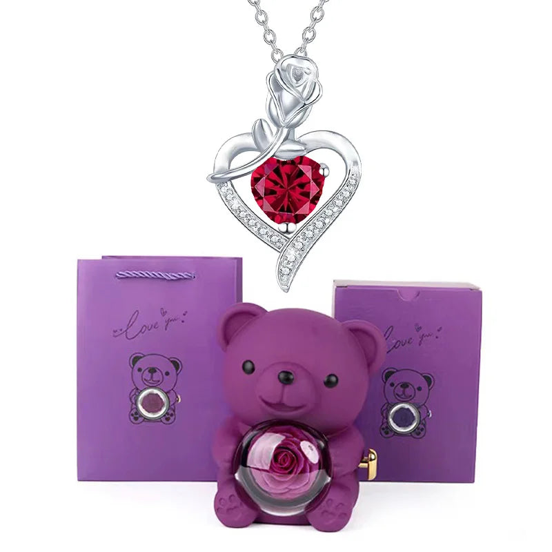 Purple teddy bear with rose, silver necklace with red heart pendant, and purple packaging on white background