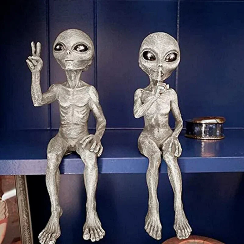 Two silver alien figurines on a blue shelf against a dark blue wall.