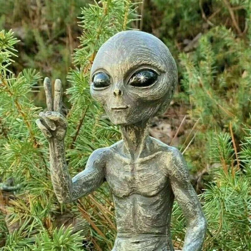 Alien figure making a peace sign gesture in front of green foliage