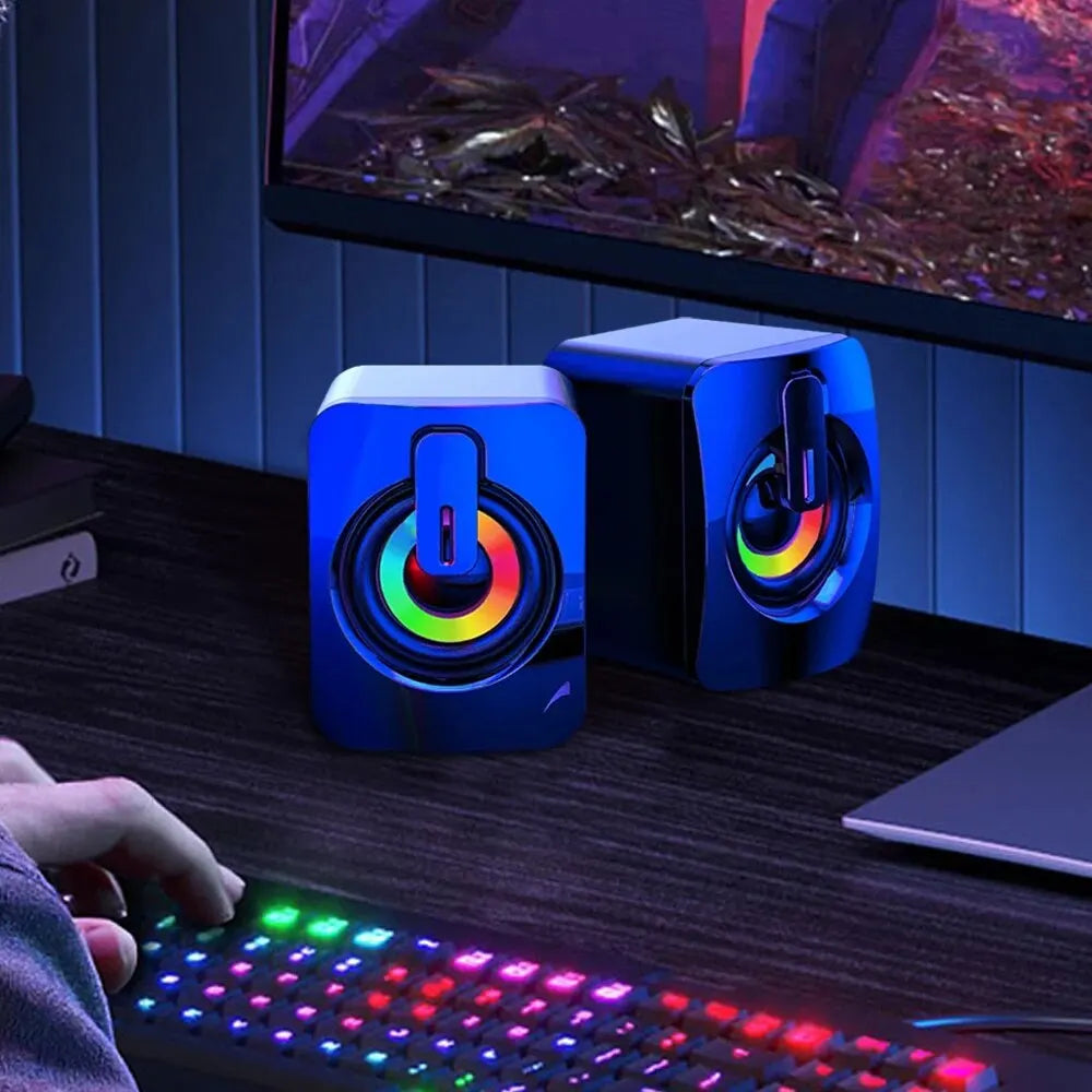 Blue gaming speakers with colorful LED lights on a desk next to a computer monitor and keyboard.