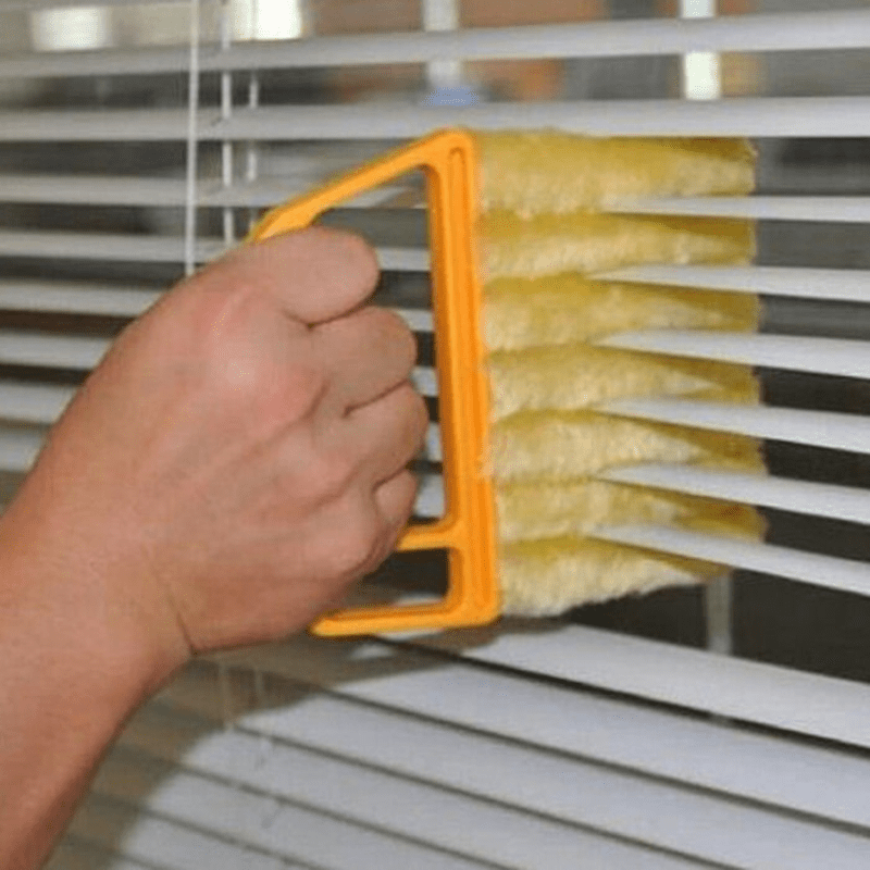 Hand using a yellow brush with an orange handle to clean blinds.