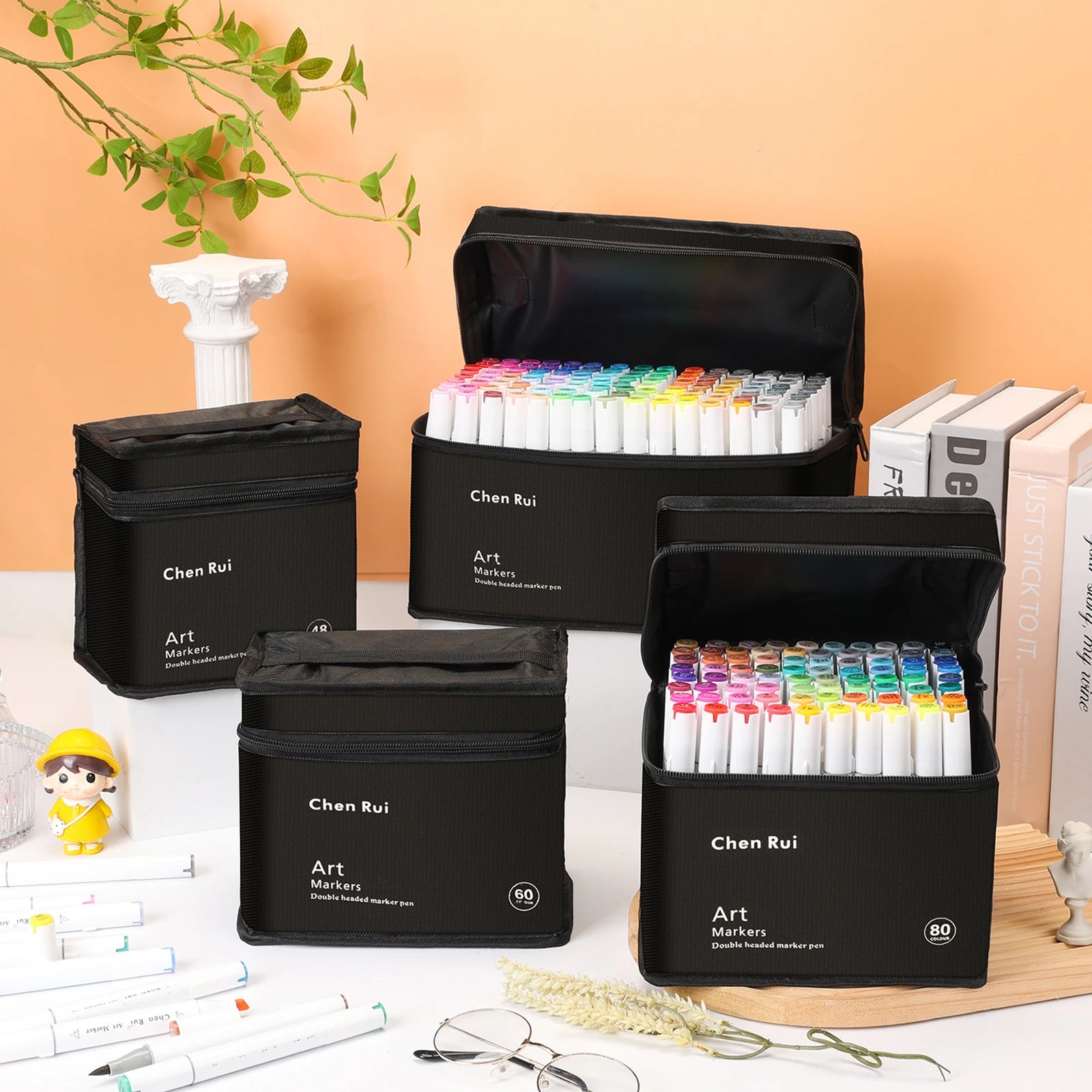 Set of Chen Rui art markers in black cases on a desk with books and a plant in the background.