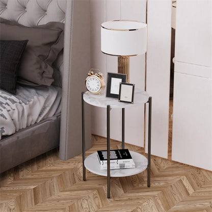 Small marble side table with a lamp, clock, and books next to a bed.