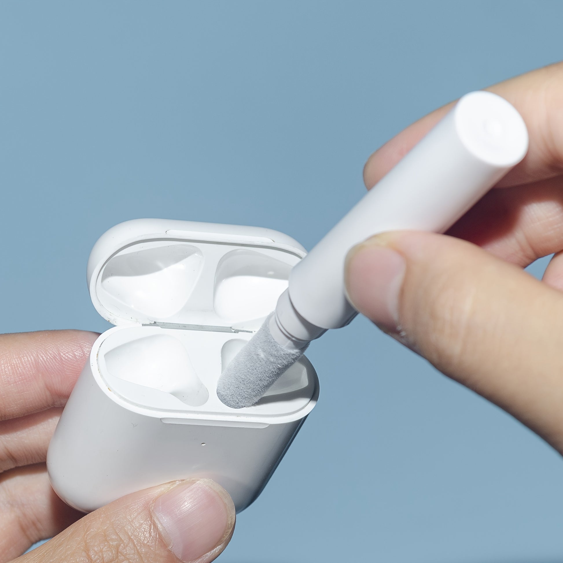 Person cleaning white earbuds with a small brush against a light blue background