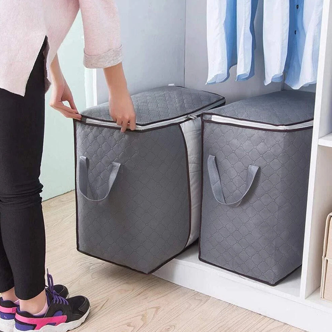 Person holding a gray quilted storage bag next to another similar bag in a room with clothes on a rack.