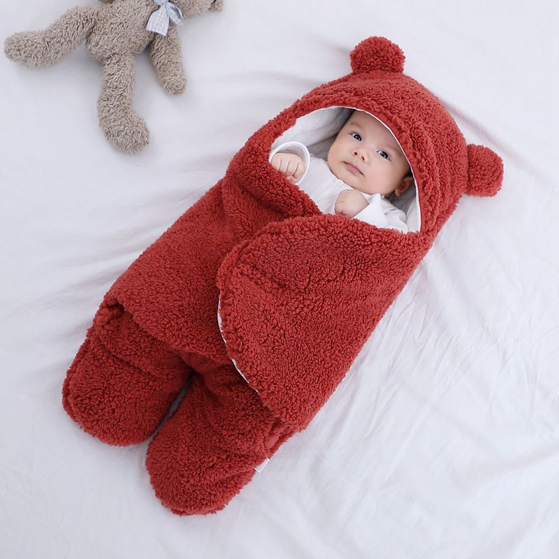 Baby in a red bear-shaped sleeping bag on a white surface with a teddy bear toy.