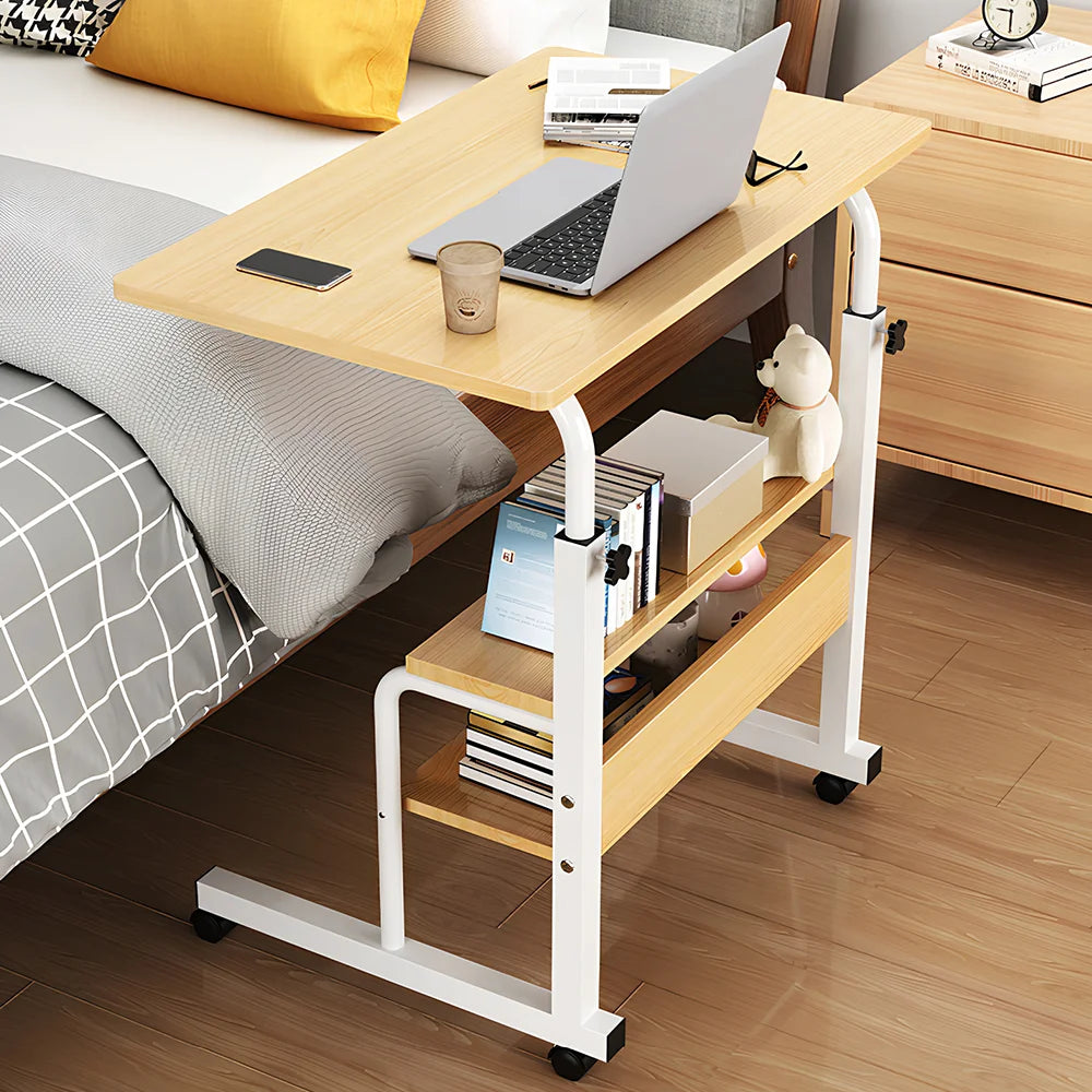 Portable desk with laptop and items on a wooden floor next to a bed.