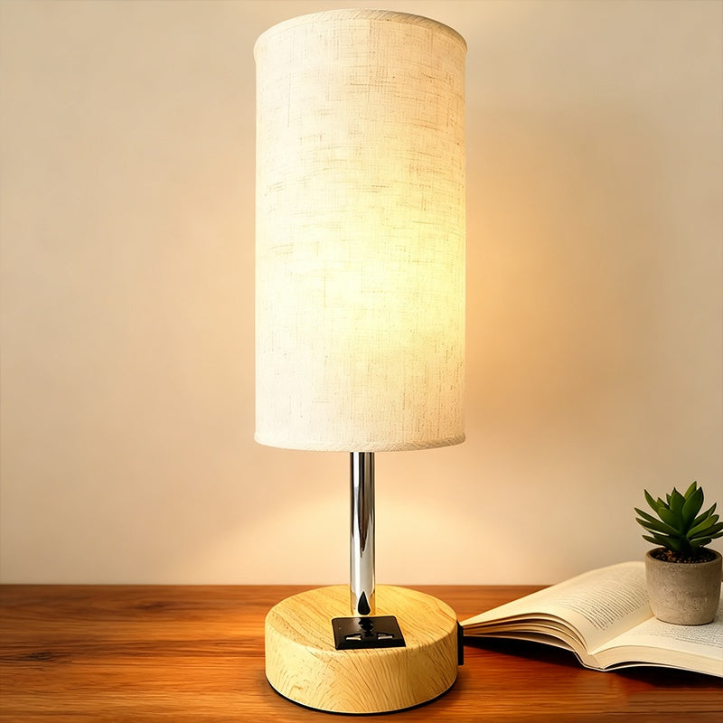 Table lamp with wooden base and white shade on a wooden surface with an open book and small plant.