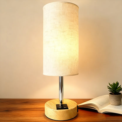 Table lamp with wooden base and white shade on a wooden surface with an open book and small plant.