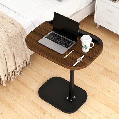 Wooden side table with laptop, mug, and watch on a wooden floor next to a bed.