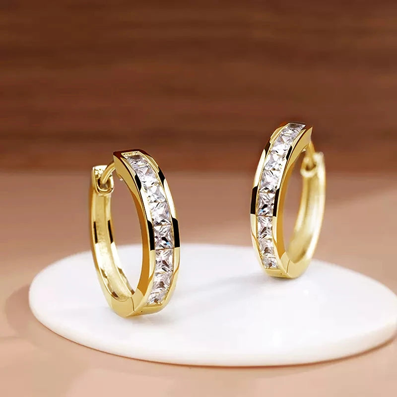 Gold hoop earrings with clear stones on a white stand against a brown background