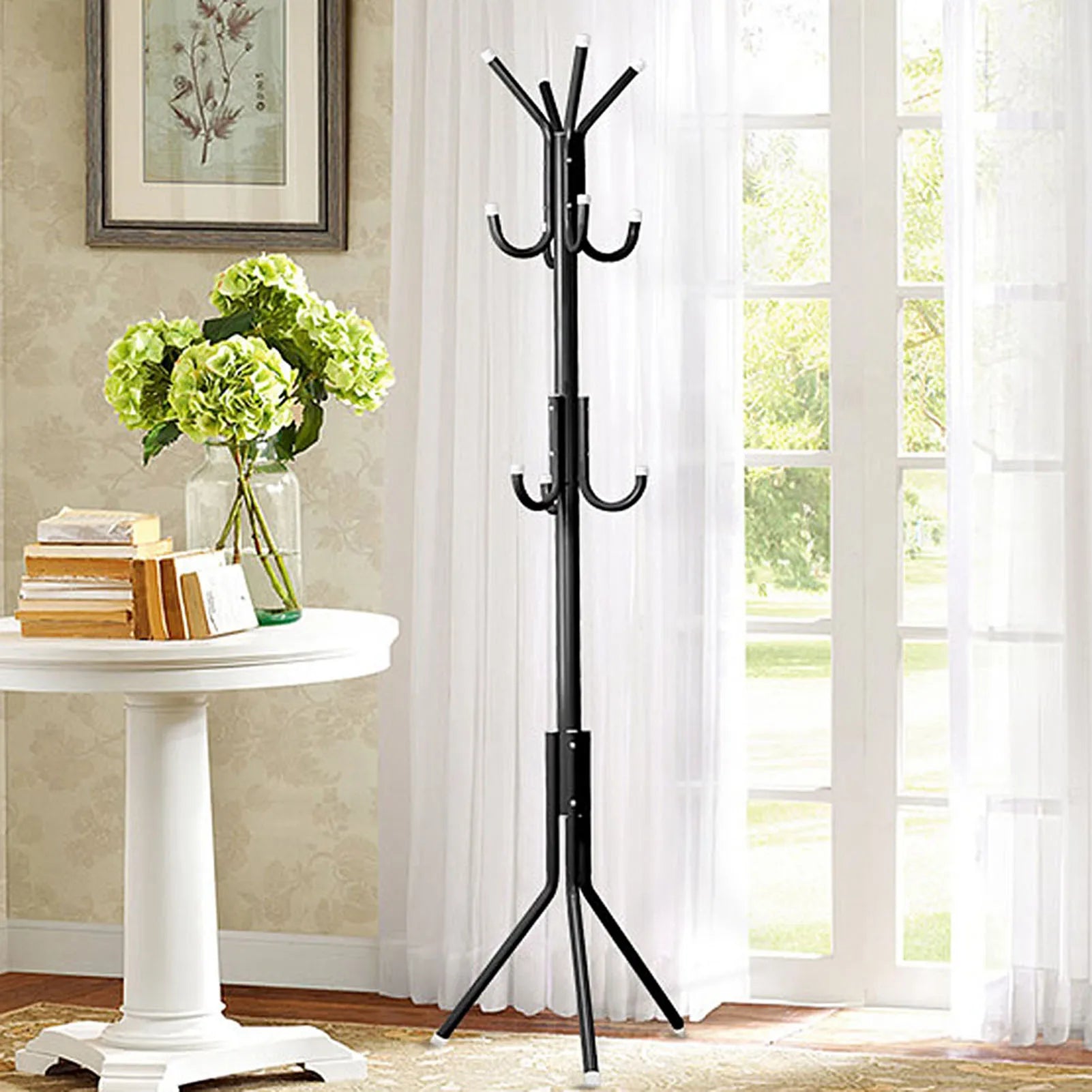 Black coat rack next to a white table with books and flowers in a room with a window and wall art.