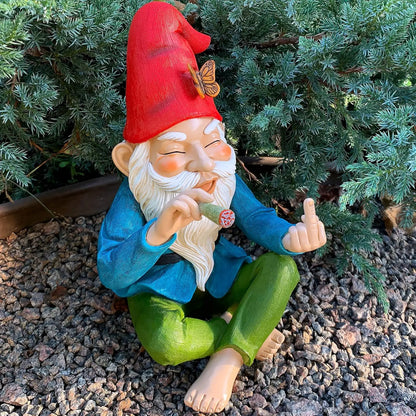 Gnome figurine with a red hat and green pants sitting on gravel with plants in the background