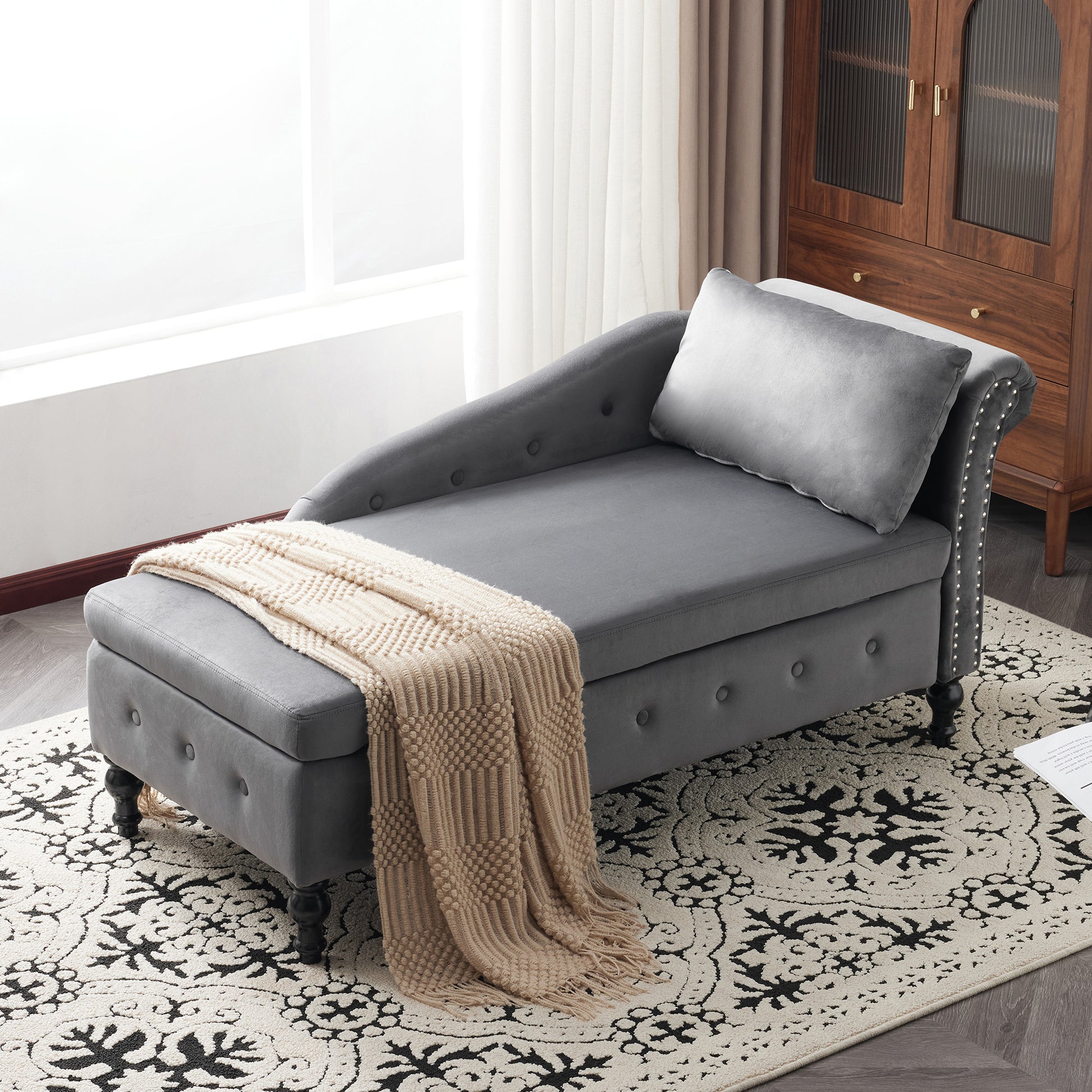 Gray tufted chaise lounge with a beige blanket on a patterned rug in a living room.