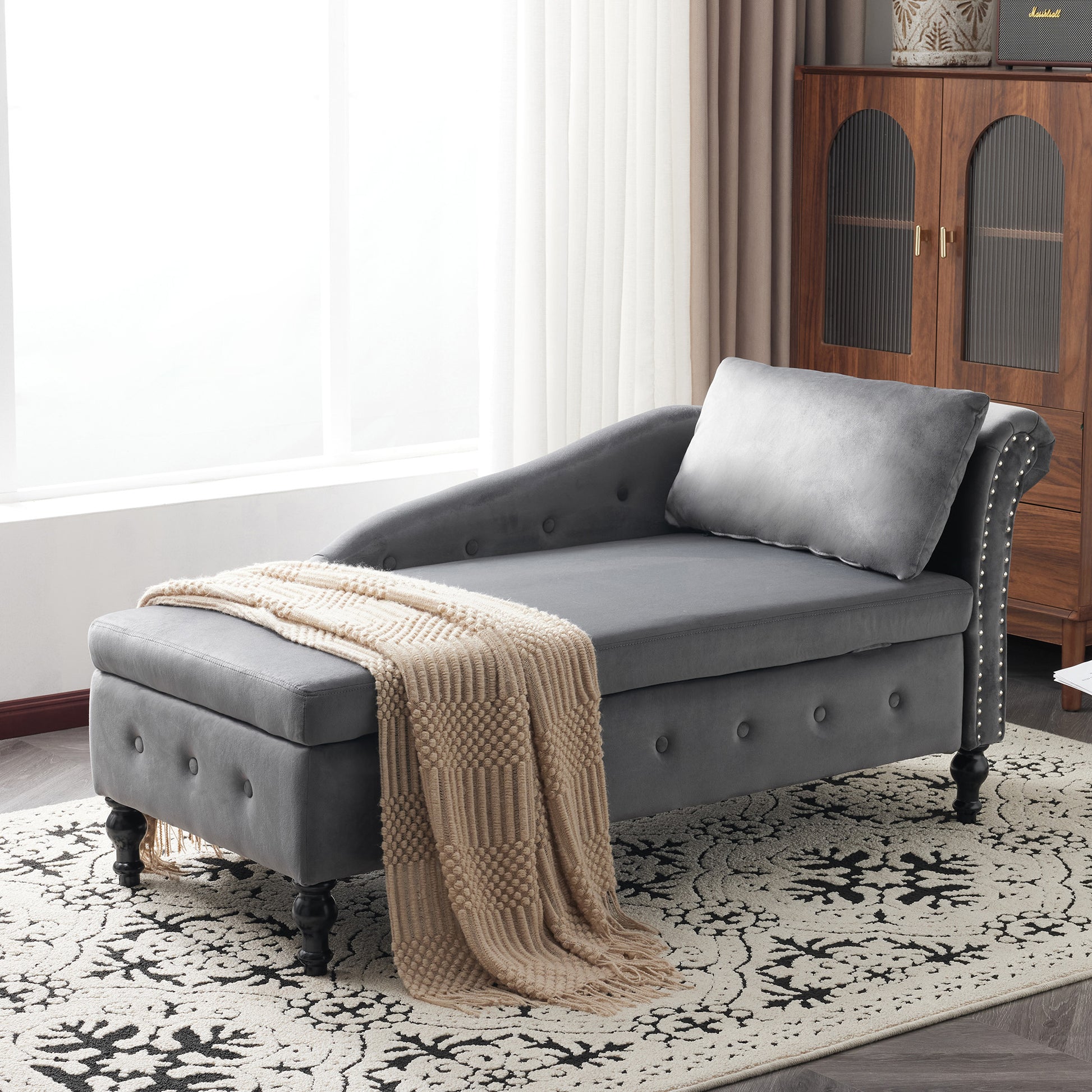 Gray tufted chaise lounge with a beige blanket on a patterned rug in a room with a wooden cabinet.