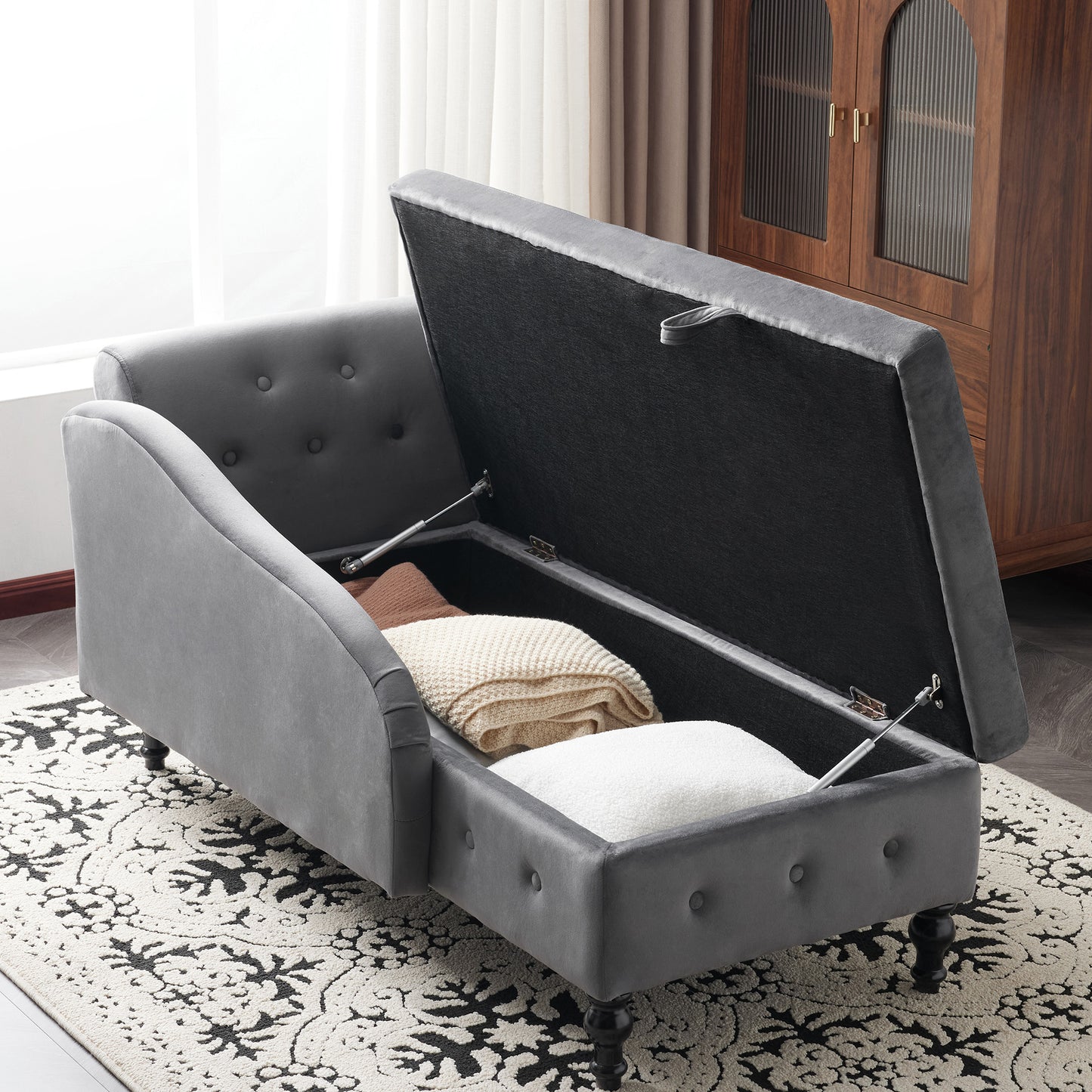 Gray upholstered storage bench with open lid revealing cushions and blankets on a patterned rug.