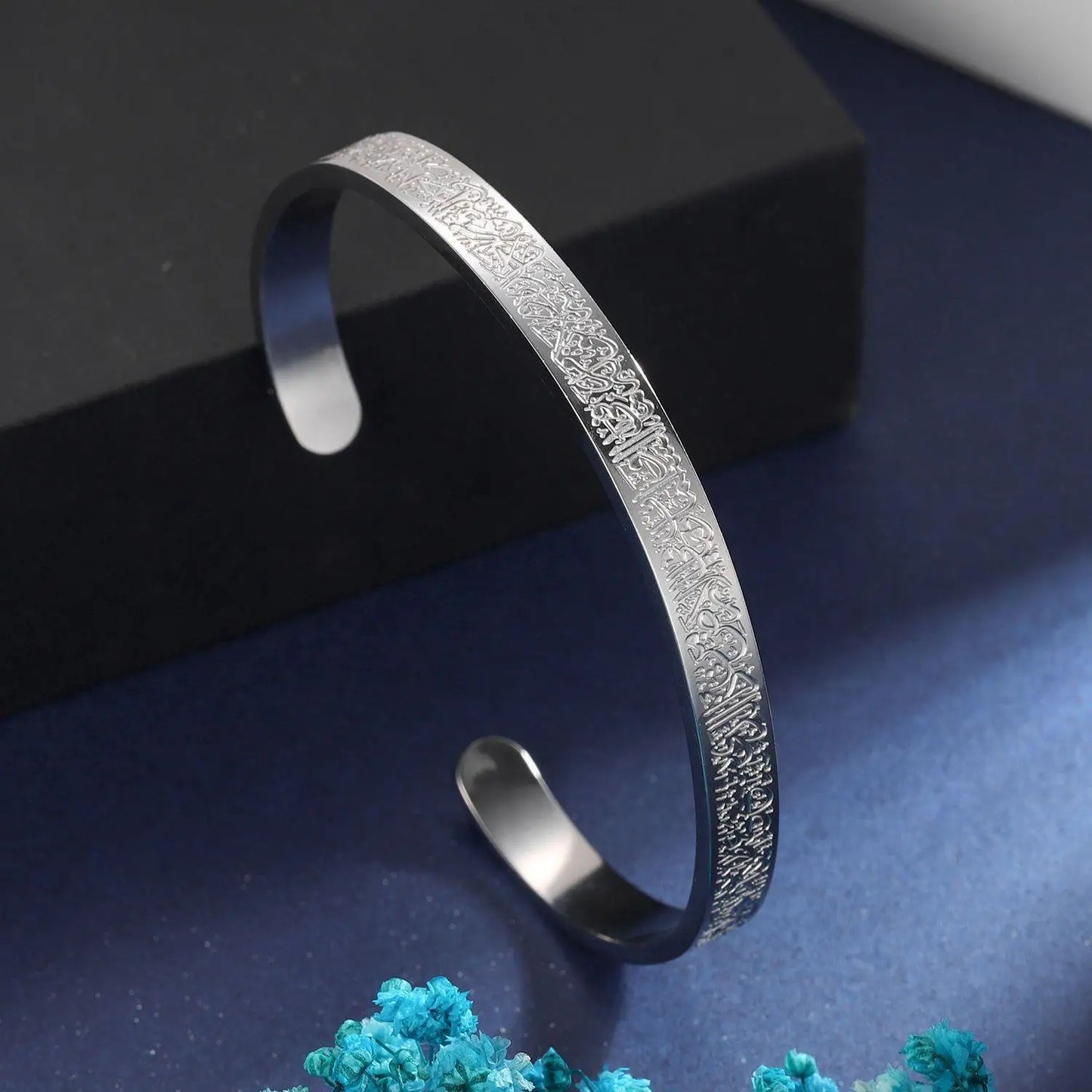 Silver bracelet with textured design on a dark blue surface with teal flowers.