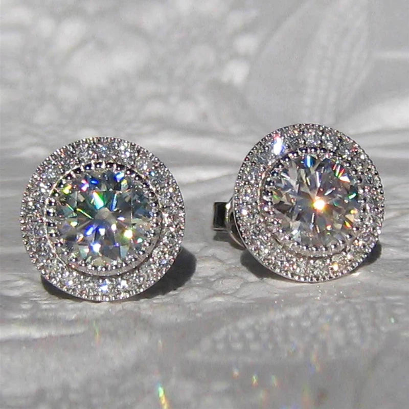 Pair of silver stud earrings with clear gemstones on a reflective surface.