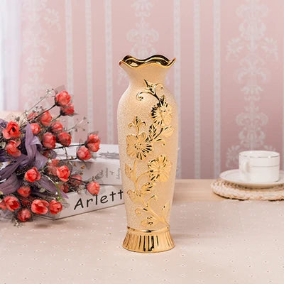 Decorative gold vase with floral patterns on a table with flowers and a box in the background.
