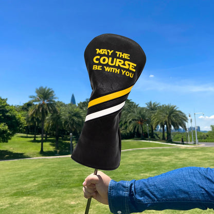 Golf club head cover with 'May the Course Be With You' text held by a person on a golf course.