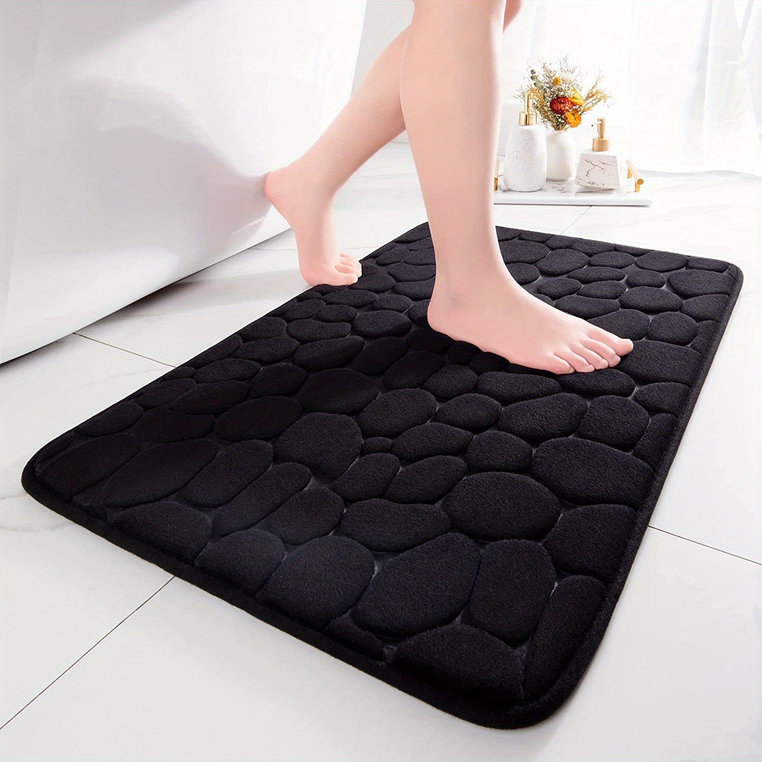 Black textured bath mat with feet stepping onto it on a white tiled floor.