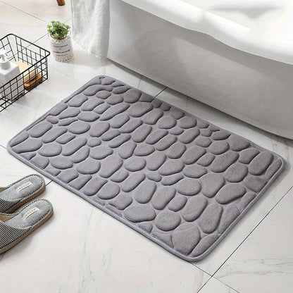 Gray textured bath mat on a bathroom floor with slippers and a plant in the background.