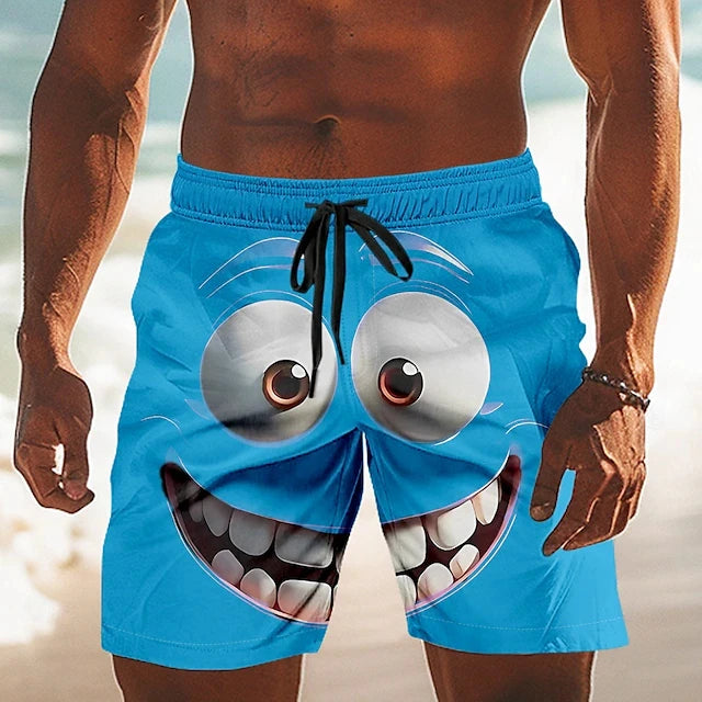 A close-up of a man wearing bright blue swim trunks with a large cartoon monster face graphic on the front, standing on a beach.