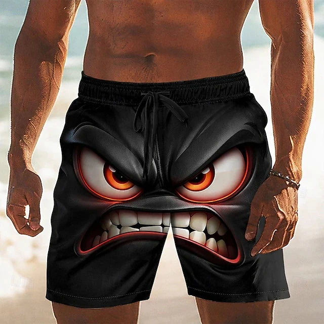 A man wearing black swim trunks with a large printed angry cartoon face design, shown from the chest down on a beach background.