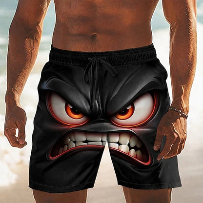 A man wearing black swim trunks with a large printed angry cartoon face design, shown from the chest down on a beach background.