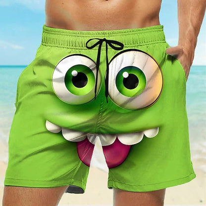 Bright green men's swim trunks with a cartoon monster face graphic, including large green eyes, white teeth, and a pink tongue, shown on a model at the beach.