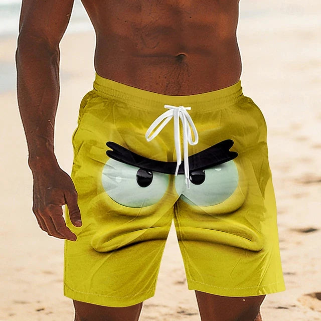 A man wearing bright yellow swim trunks with a large cartoon angry-eye graphic on the front, standing on a sandy beach.