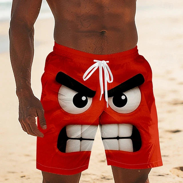 A close-up of a person wearing bright red swim trunks with a cartoon angry face graphic on the front, standing on a sandy beach.