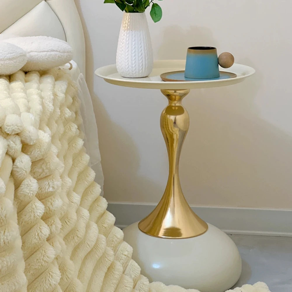 Small round table with gold base and white top next to a textured white blanket on a bed.